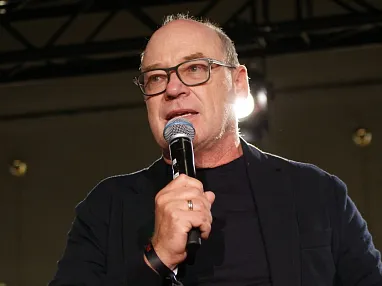 Mann mit Brille und Mikrofon spricht auf einer Bühne, trägt ein dunkles Jackett, dunkles Hemd und steht vor einem beleuchteten Hintergrund.