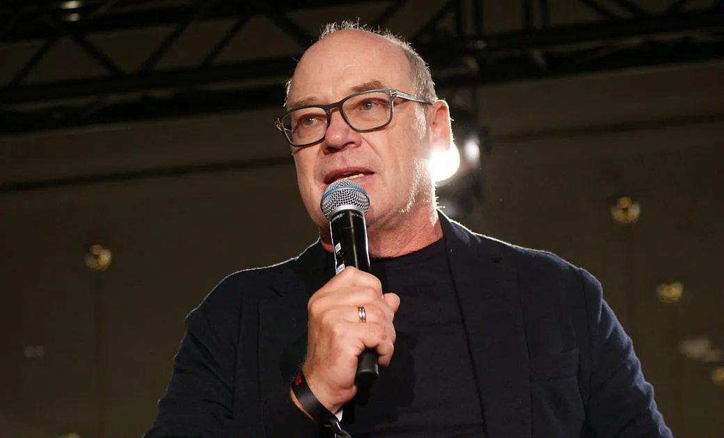 Mann mit Brille und Mikrofon spricht auf einer Bühne, trägt ein dunkles Jackett, dunkles Hemd und steht vor einem beleuchteten Hintergrund.