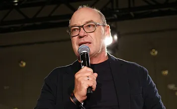 Mann mit Brille und Mikrofon spricht auf einer Bühne, trägt ein dunkles Jackett, dunkles Hemd und steht vor einem beleuchteten Hintergrund.