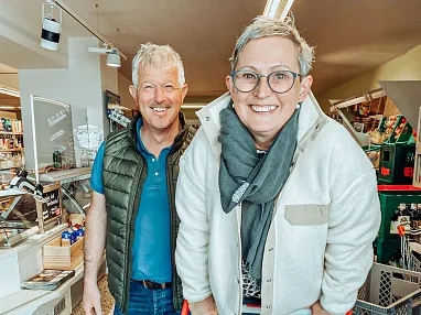 Ein Paar lächelt in einem Supermarkt. Die Frau stützt sich auf einen Einkaufswagen, während der Mann neben ihr steht. Beide wirken fröhlich und entspannt.