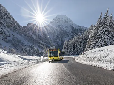 Ein gelber Bus fährt auf einer verschneiten Bergstraße, umgeben von schneebedeckten Tannen und majestätischen Bergen. Die Sonne scheint hell über dem Gipfel.