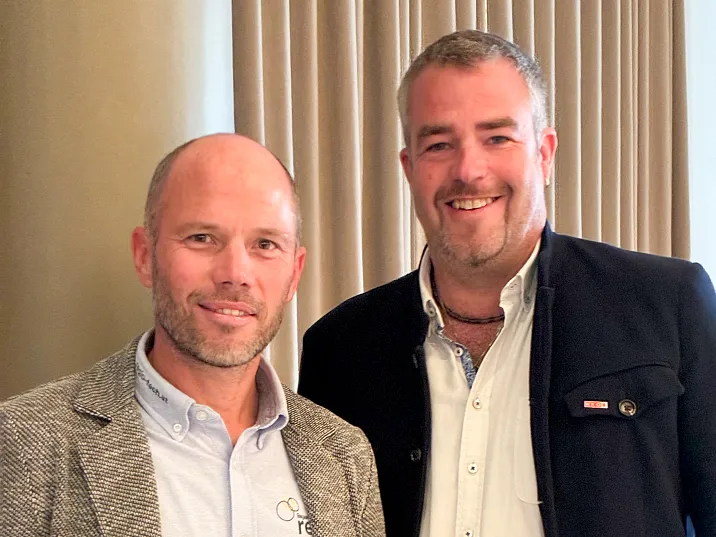 Zwei Männer in formeller Kleidung stehen nebeneinander vor beigen Vorhängen. Sie lächeln in die Kamera. Einer trägt ein Sakko, der andere eine dunkle Jacke.