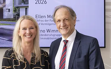 Ein Mann und eine Frau in Business-Kleidung lächeln in die Kamera. Im Hintergrund ist ein Poster mit Text und einem modernen Gebäude sichtbar.