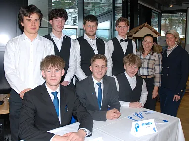 Eine Gruppe junger Menschen in formeller Kleidung sitzt und steht an einem Tisch. Zwei Frauen in Freizeitkleidung stehen rechts daneben.