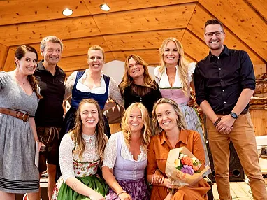 Eine fröhliche Gruppe von Menschen in traditioneller bayerischer Tracht posiert auf einer hölzernen Bühne vor einem rustikalen Hintergrund.