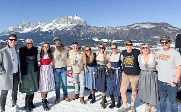 Gruppe von Menschen in traditioneller Tracht steht lächelnd auf schneebedecktem Boden vor malerischer Berglandschaft und blauem Himmel.