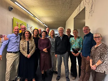 Eine Gruppe von zwölf lächelnden Personen steht nebeneinander in einem Flur mit moderner Dekoration und natürlichem Licht.