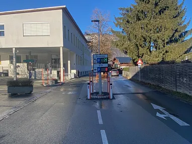 Eine leere Straße bei Sonnenschein mit einer Schranke, einem Gebäude links und Bäumen im Hintergrund. Verkehrszeichen sind aufgestellt.