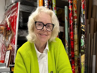 Eine lächelnde ältere Frau mit blonder Frisur und großer Brille trägt einen leuchtend grünen Cardigan vor einer Wand mit bunten Geschenkpapierrollen.