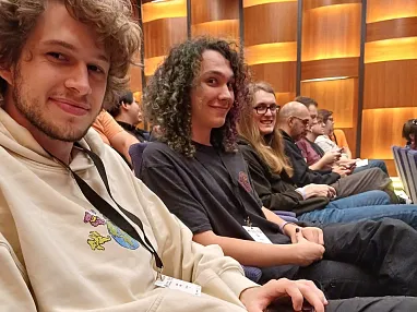 Gruppe von Menschen sitzt in einem Auditorium, lächelnd, einige mit Namensschildern. Der Raum hat eine holzgetäfelte Wand im Hintergrund.