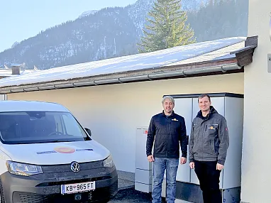 Zwei Männer stehen lächelnd vor einem weißen Lieferwagen und einem Gebäude mit schneebedecktem Dach. Im Hintergrund sind verschneite Berge zu sehen.