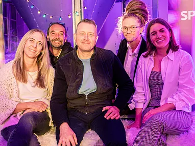 Fünf Personen sitzen lächelnd zusammen in einem gemütlichen, bunt beleuchteten Raum. Sie wirken entspannt und fröhlich, umgeben von dekorativen Lichtern.