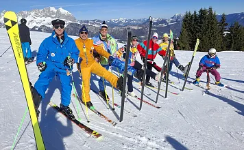 Eine Gruppe von Skifahrern in bunter Kleidung posiert fröhlich auf einer verschneiten Piste mit Bergen im Hintergrund und strahlend blauem Himmel.