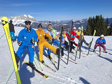 Eine Gruppe von Skifahrern in bunter Kleidung posiert fröhlich auf einer verschneiten Piste mit Bergen im Hintergrund und strahlend blauem Himmel.