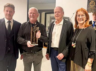 Vier Personen stehen nebeneinander, eine Person hält eine Trophäe. Alle sind schick gekleidet und wirken feierlich. Im Hintergrund ein Kellner mit Getränken.