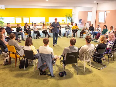 Person steht in der Mitte eines Stuhlkreises und spricht zu einer Gruppe von Menschen in einem hellen Raum mit gelben Wänden und großen Fenstern.