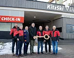Mehrere Personen in roten Jacken stehen zusammen vor einem Gebäude und halten einen großen symbolischen Schlüssel. Im Hintergrund sind "Self CHECK-IN" Schilder zu sehen.