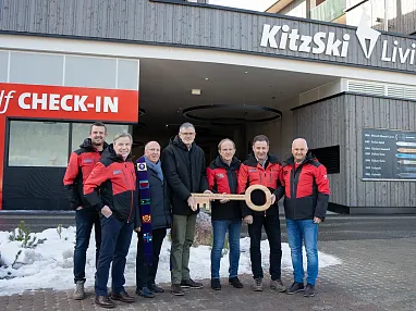 Mehrere Personen in roten Jacken stehen zusammen vor einem Gebäude und halten einen großen symbolischen Schlüssel. Im Hintergrund sind "Self CHECK-IN" Schilder zu sehen.