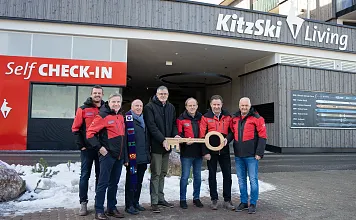 Mehrere Personen in roten Jacken stehen zusammen vor einem Gebäude und halten einen großen symbolischen Schlüssel. Im Hintergrund sind "Self CHECK-IN" Schilder zu sehen.