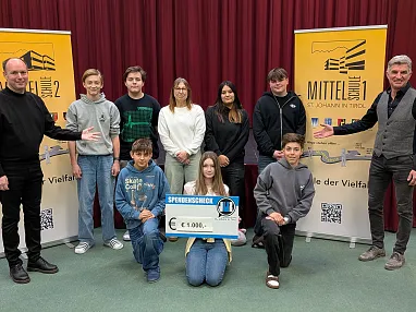 Eine Gruppe von Jugendlichen und zwei Erwachsenen posieren auf einer Bühne mit einem großen Spendenscheck vor Aufstellern mit dem Text "Mittel der Vielfalt".