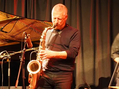 Ein Musiker spielt Saxophon auf einer Bühne mit schwarzem Vorhang, umgeben von verschiedenen Instrumenten. Die Beleuchtung ist warm und gemütlich.