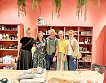 Eine Gruppe von fünf Menschen steht lächelnd in einem gemütlichen Laden mit roten Wänden, dekorativen Pflanzen und Regalen voller Produkte.