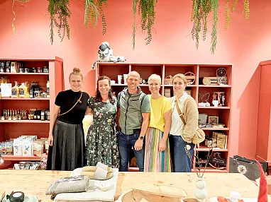 Eine Gruppe von fünf Menschen steht lächelnd in einem gemütlichen Laden mit roten Wänden, dekorativen Pflanzen und Regalen voller Produkte.