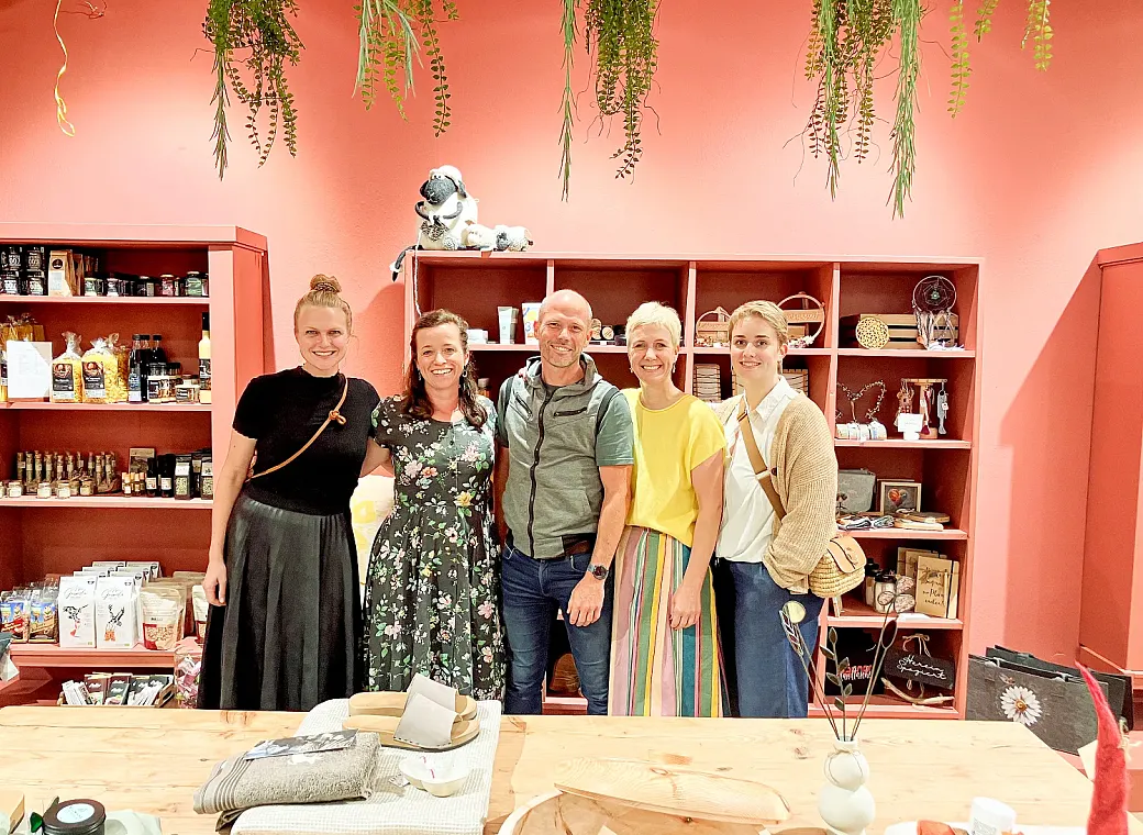 Eine Gruppe von fünf Menschen steht lächelnd in einem gemütlichen Laden mit roten Wänden, dekorativen Pflanzen und Regalen voller Produkte.