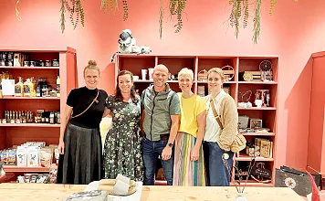 Eine Gruppe von fünf Menschen steht lächelnd in einem gemütlichen Laden mit roten Wänden, dekorativen Pflanzen und Regalen voller Produkte.