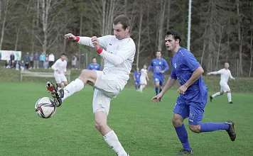 Zwei Fußballspieler während eines Spiels: Ein Spieler in Weiß kontrolliert den Ball, während ein Spieler in Blau hinter ihm herläuft. Grüne Spielfläche.