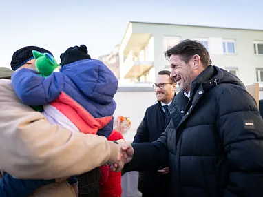 Ein Mann in einer Winterjacke schüttelt die Hand eines anderen Mannes, der ein Kind hält. Beide lächeln, während sie sich unter freiem Himmel treffen.