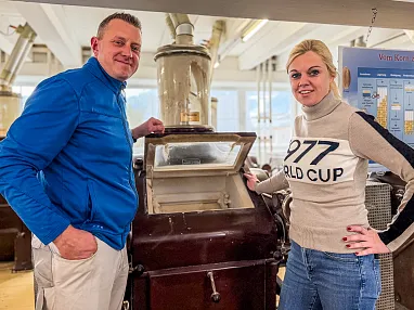 Zwei Personen stehen in einer historischen Mühle. Sie lächeln und zeigen auf eine alte Maschine. Im Hintergrund sind Schilder und große Fenster sichtbar.