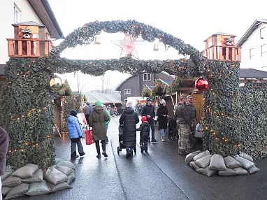 Besucher schlendern durch einen festlich geschmückten Weihnachtsmarkt mit Lichterketten, Tannengirlanden und dekorierten Ständen in winterlicher Atmosphäre.
