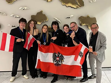 Eine Gruppe von sieben Personen posiert mit österreichischen Flaggen und Medaillen, lächelnd vor einer weißen Wand mit goldenen Verzierungen.