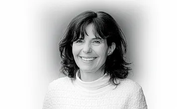 Porträtfoto einer lächelnden Frau mit welligem, dunklem Haar in einem weißen Oberteil vor einem neutralen Hintergrund in Schwarz-Weiß.