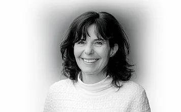 Porträtfoto einer lächelnden Frau mit welligem, dunklem Haar in einem weißen Oberteil vor einem neutralen Hintergrund in Schwarz-Weiß.