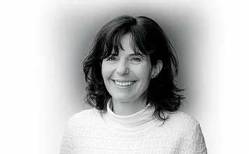 Porträtfoto einer lächelnden Frau mit welligem, dunklem Haar in einem weißen Oberteil vor einem neutralen Hintergrund in Schwarz-Weiß.