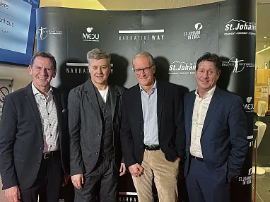 Vier Männer in Anzügen stehen lächelnd vor einer bedruckten Wand mit Logos und Schriftzügen bei einem Event.