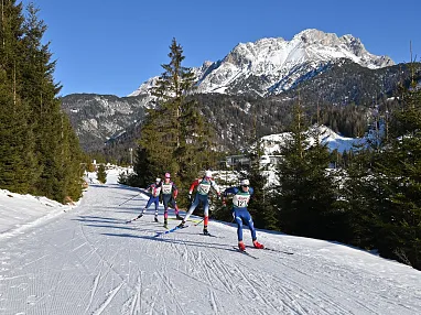 Langläufer in bunten Anzügen auf einer verschneiten Loipe, umgeben von Nadelbäumen und Bergen im Hintergrund unter einem klaren blauen Himmel.