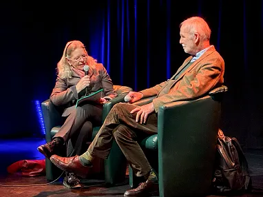 Zwei Personen sitzen auf einer Bühne in grünen Sesseln, eine Frau hält ein Mikrofon und spricht mit einem Mann. Der Hintergrund ist dunkel mit blauer Beleuchtung.