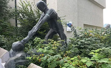 Skulptur eines stilisierten Menschen, der sich über einen rechteckigen Steinsockel beugt, umgeben von üppigem Grün in einem urbanen Umfeld.