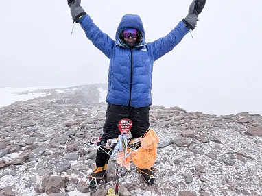 Person in blauer Jacke steht im Schnee auf einem Berggipfel, hebt triumphierend die Arme und trägt Kletterausrüstung. Der Himmel ist bewölkt.