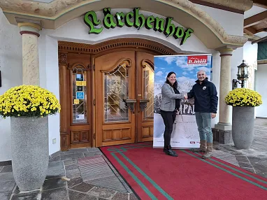 Zwei Personen stehen vor einem Gebäude mit der Aufschrift "Lärchenhof", lächelnd und Händeschütteln. Neben ihnen blühen gelbe Chrysanthemen in großen Töpfen.