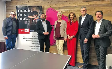 Sechs Personen stehen in einem Büro vor einer Holzvertäfelung und einem Banner mit der Aufschrift "macht.mir.mut". Ein Tisch mit pinkfarbenem Detail ist sichtbar.