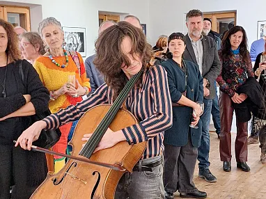 Eine Cellistin spielt leidenschaftlich ihr Instrument, umgeben von einem Publikum in einem Kunstgalerie-Ambiente. Menschen zeigen Interesse und hören zu.