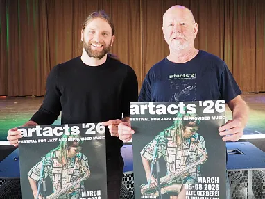 Zwei Männer stehen auf einer Bühne und halten Plakate für das Artacts '26 Festival vor sich. Der Hintergrund zeigt Vorhänge und Lautsprecher.