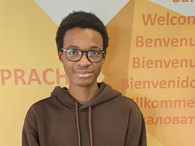 Ein junger Mensch mit Brille und braunem Kapuzenpullover steht vor einer Wand mit Begrüßungen in verschiedenen Sprachen.