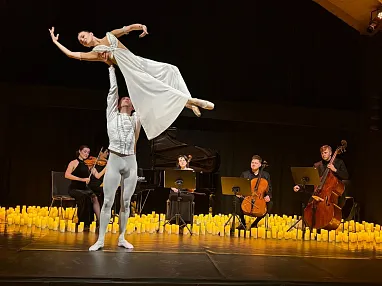 Ein Ballettpaar tanzt auf einer Bühne, umgeben von vielen Kerzen. Im Hintergrund spielen Musiker auf Klavier, Cello und Kontrabass. Elegante und stimmungsvolle Szene.