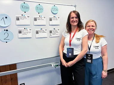 Zwei Frauen stehen vor einem Whiteboard mit Zeitplänen und lächeln in die Kamera. Beide tragen weiße T-Shirts und Namensschilder um den Hals.