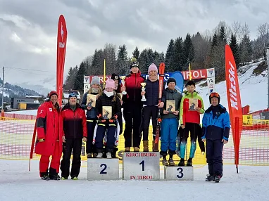 Eine Gruppe von Menschen steht auf einem Podest im Schnee. Alle tragen Winterkleidung. Drei Personen halten Auszeichnungen und lächeln in die Kamera.